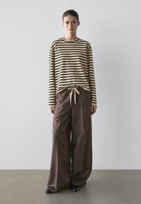 Giovane donna in piedi che indossa una maglia a maniche lunghe a righe beige e nere con pantaloni larghi marroni legati in vita e scarpe nere, sfondo neutro.