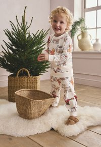 Children's pajama set in cream with colorful holiday-themed prints. Soft fabric, ribbed cuffs, and matching slippers. Cozy setting with a tree.