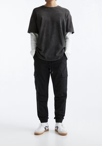 Schwarzes T-Shirt mit kurzen Ärmeln, über einem weißen Langarmshirt getragen, kombiniert mit schwarzen Cargo-Hosen und weißen Sneakers mit braunen Sohlen.