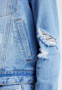 Ljusblå denimjacka med en stor rivning på ärmen, fransiga kanter, en bröstficka och synliga sömnadsdetaljer.