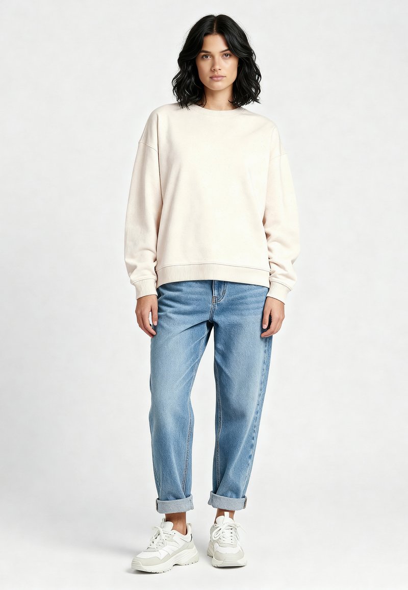 Sweatshirt beige chiaro con maniche lunghe e scollatura arrotondata, abbinato a jeans azzurri arrotolati e sneakers bianche.