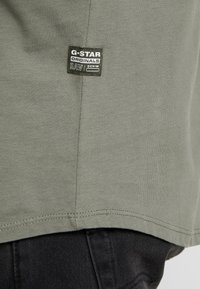 Malá černá nášivka na lemu khaki košile, na které je tekst "G-STAR ORIGINALS RAW DENIM", pod kterou se nacházejí černé džínové kalhoty.