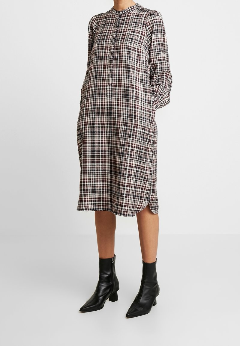 Femme portant une robe à carreaux à manches longues, longueur genou, avec un col mandarin et des bottines noires, debout devant un fond blanc.