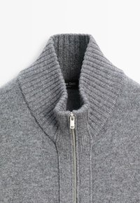 Maglione grigio lavorato a maglia con colletto alto a coste, dotato di una chiusura con zipper argentata e realizzato in un tessuto morbido e testurizzato. Etichetta del marchio visibile all'interno del colletto.