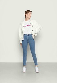 Casaco branco sobre uma t-shirt gráfica com "Magic Monda" em rosa, combinado com jeans skinny azul claro e ténis brancos. Fundo simples.