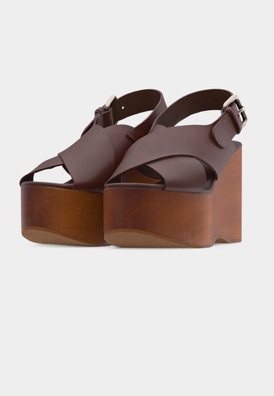 Sandales compensées en cuir marron avec larges brides croisées, boucle à la cheville, et talons compensés épais en bois sur fond blanc.