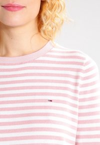 Randig tröja i rosa och vitt, med ribbat halslinning och liten logotypdetalj på bröstet. Mjuk stickad textur, avslappnad passform.