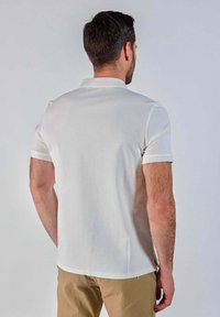 Homme aux cheveux courts et sombres portant un polo blanc à manches courtes et un pantalon beige, se tenant de dos contre un fond uni.