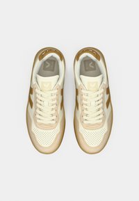 Ζεύγος μπεζ και λευκά αθλητικά παπούτσια Veja με καουτσούκ σόλες και καφέ λογότυπα "V", απεικονιζόμενα από ψηλά σε λευκό φόντο.