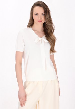 Witte top met korte mouwen en een V-hals, voorzien van een strikdetail bij de kraag. Gladde textuur, aansluitende stijl, gecombineerd met luchtige lichtgekleurde broeken.