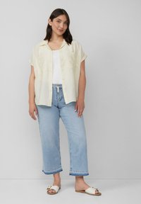 Lichtgele overhemd met korte mouwen, knoopsluiting en twee borstzakken, gedragen over een witte tanktop, gecombineerd met lichtblauwe wijde jeans en witte sandalen.