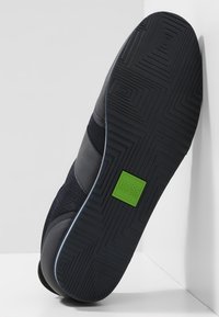 Svart sneakers med överdel i nät och läder, med en texturerad gummisul, grön logotypdetalj och en lågprofildesign.