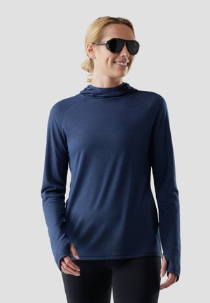 Kobieta w czarnych okularach przeciwsłonecznych i niebieskiej, teksturowanej bluzie z długim rękawem z otworami na kciuki, uśmiechająca się na jednolitym jasnym tle.