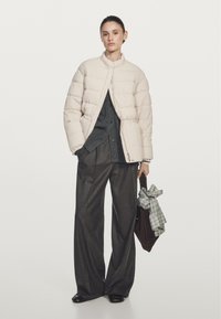 Donna con un giubbotto imbottito beige sopra una camicia scura a bottoni e pantaloni a gamba ampia, che tiene una borsa marrone con un panno a quadri annodato.