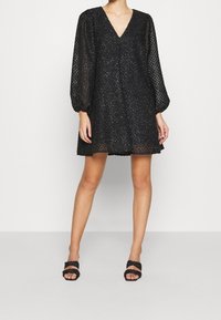 Vestido negro corto con escote en V, mangas abullonadas y patrón texturizado. Lleva unas sandalias negras con tiras. Tela suave con un acabado ligeramente brillante.