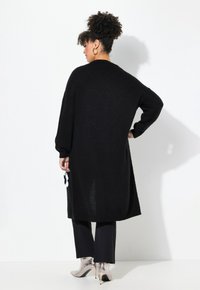 Langer schwarzer Cardigan aus weichem Strickmaterial, mit herunterfallenden Schultern, gerippten Säumen und einem dekorativen weißen Blumenakzent an der Seite.