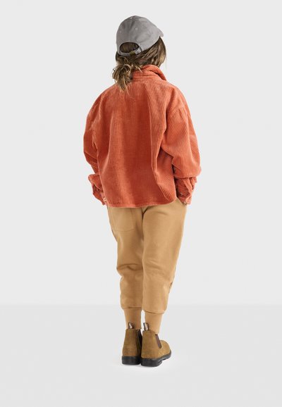 Oranžová kordurová oversized košeľa, béžové tepláky a hnedé členkové topánky. Šedá čiapka dotvára oblečenie, pohľad zozadu.