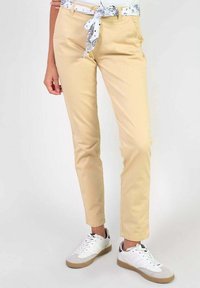 Chinos beige à coupe slim, avec des poches avant et une ceinture de tissu blanche, associés à des baskets blanches et grises.