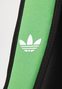Un tessuto verde con una finitura texture presenta un logo Adidas bianco e tre strisce, bordato da materiale nero.