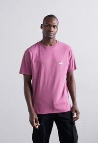 Camiseta de algodón rosa con cuello redondo y mangas cortas, que presenta un pequeño logo blanco en el pecho. Combinada con pantalones cargo negros.