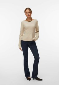 Femme debout portant un pull beige, un jean bleu foncé, des chaussures noires pointues et un collier épais à perles sur un fond blanc.