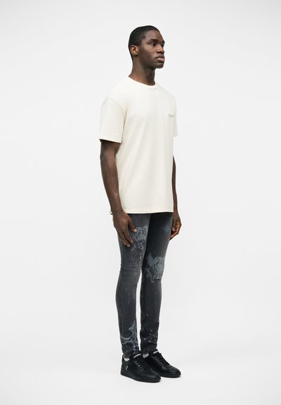 Άνδρας που στέκεται φορώντας ένα απλό, εκρού T-shirt, μαύρο τζιν με σχέδια skinny, και μαύρα αθλητικά παπούτσια, μπροστά σε λευκό φόντο.