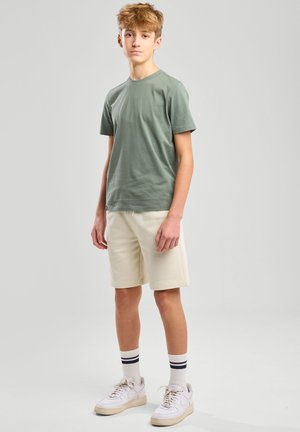 Grünes Baumwoll-T-Shirt, cremefarbene Shorts, weiße Sneakers mit hellen Akzenten und gestreifte Socken. Das Model steht vor einem schlichten grauen Hintergrund.