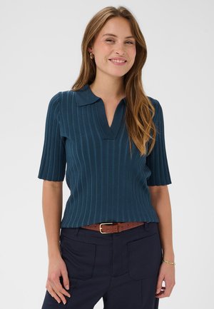 Türkiissinine soonikkoes lühikeste varrukatega polo-särk, millel on v-kaelus ja tekstuuriga kangas, kombineeritud tumedate pükste ning pruuni vöökohaga rihmaga.