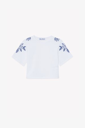 Weißes, kurzes T-Shirt mit kurzen Ärmeln, verziert mit blauer Blumenstickerei auf den Schultern. Hergestellt aus weichem Baumwollmaterial.