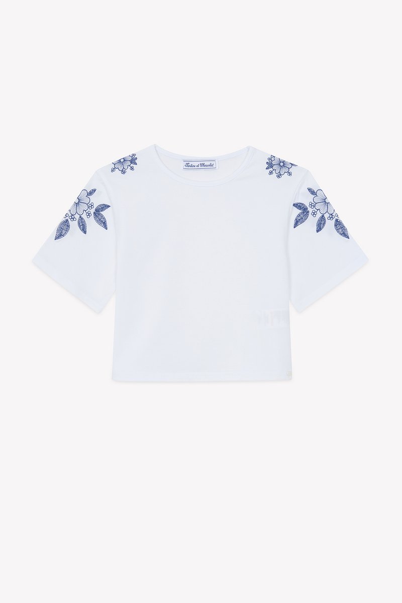 Λευκό κοντό t-shirt με κοντά μανίκια, διακοσμημένο με μπλε κεντήματα λουλουδιών στους ώμους. Κατασκευασμένο από απαλό βαμβακερό ύφασμα.