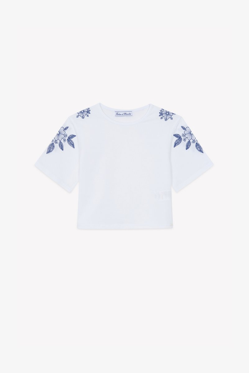 Λευκό κοντό t-shirt με κοντά μανίκια, διακοσμημένο με μπλε κεντήματα λουλουδιών στους ώμους. Κατασκευασμένο από απαλό βαμβακερό ύφασμα.