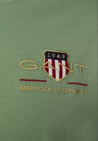 Sudadera de algodón verde con un escudo bordado que presenta rayas rojas y blancas, "GANT" en dorado y "AMERICAN SPORTSWEAR" debajo.