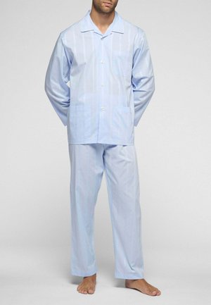 Uomo in piedi a piedi nudi che indossa una camicia da pigiama azzurro chiaro a maniche lunghe con bottoni e pantaloni da pigiama larghi abbinati su sfondo semplice.
