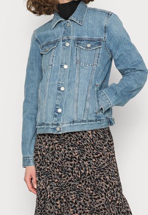 Osoba nosząca jasnoniebieską kurtkę dżinsową na czarnym topie z plisowaną spódnicą w czarno-brązowo-beżowy wzór, stojąca na jednolitym tle.