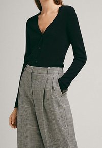 Cardigan noir à col en V avec fermeture à boutons, associé à un pantalon gris à carreaux taille haute avec plis et poches latérales.