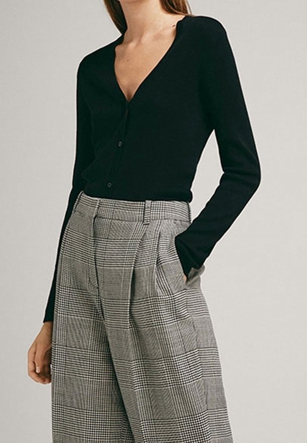 Cardigan noir à col en V avec fermeture à boutons, associé à un pantalon gris à carreaux taille haute avec plis et poches latérales.