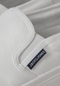 Fehér vászon slip-on cipő, texturált felülettel, steppelt szélekkel és egy fekete címkével, amin fehér betűkkel szerepel a "JACK & JONES" felirat.