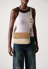 Woven beige and tan crossbody bag with a flap, featuring a light strap and subtle stitching details. Model wearing a pink tank top and denim.