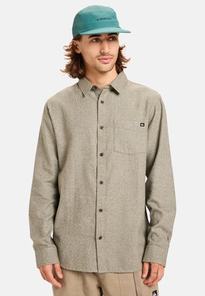 Flannel shirt in light brown with a chest pocket, button closure, and long sleeves. Paired with a teal cap featuring a logo detail.