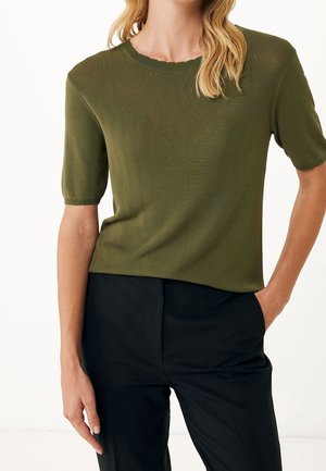 Frau trägt ein olivgrünes, kurzärmeliges Strickoberteil in Kombination mit hoch taillierten schwarzen Hosen, steht mit einer Hand in der Hosentasche.