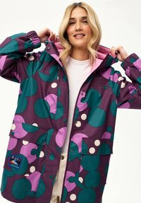 Uśmiechnięta blondynka poprawiająca kaptur fioletowej kurtki z dużymi zielonymi, różowymi i białymi okrągłymi wzorami, ubrana w białą koszulę i beżowe spodnie.