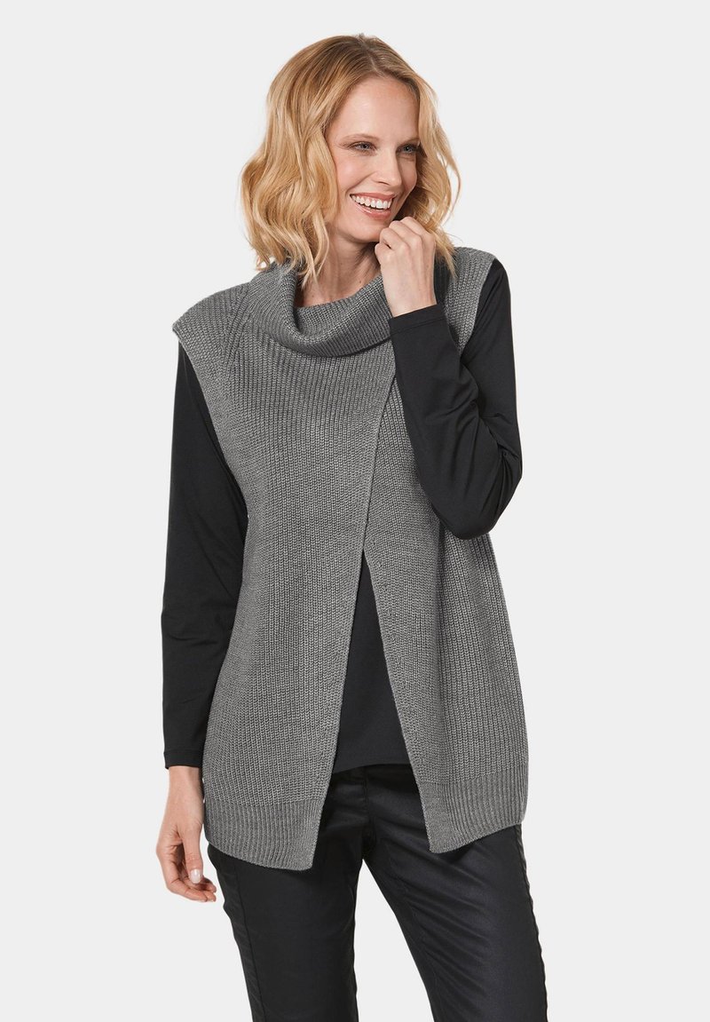 Grauer gestrickter ärmelloser Pullover mit einem Schalkragen, der ein strukturiertes Design und eine offene Vorderseite aufweist, über einem schwarzen Langarmshirt getragen.