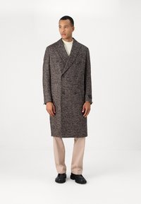 Abrigo de lana texturizada marrón y cream de dos filas de botones, con mangas largas, bolsillos laterales y solapa tipo muesca, llevado sobre un suéter cream y pantalones claros.