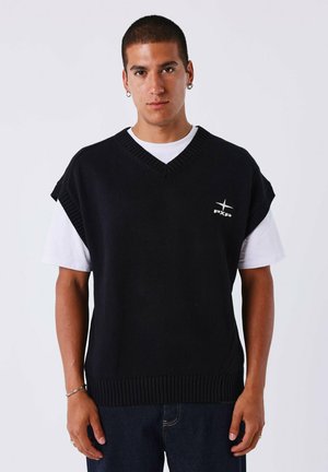 Gilet noir en tricot avec un col en V et de courtes manches droites. Logo blanc sur la poitrine. Porté par-dessus un T-shirt blanc.