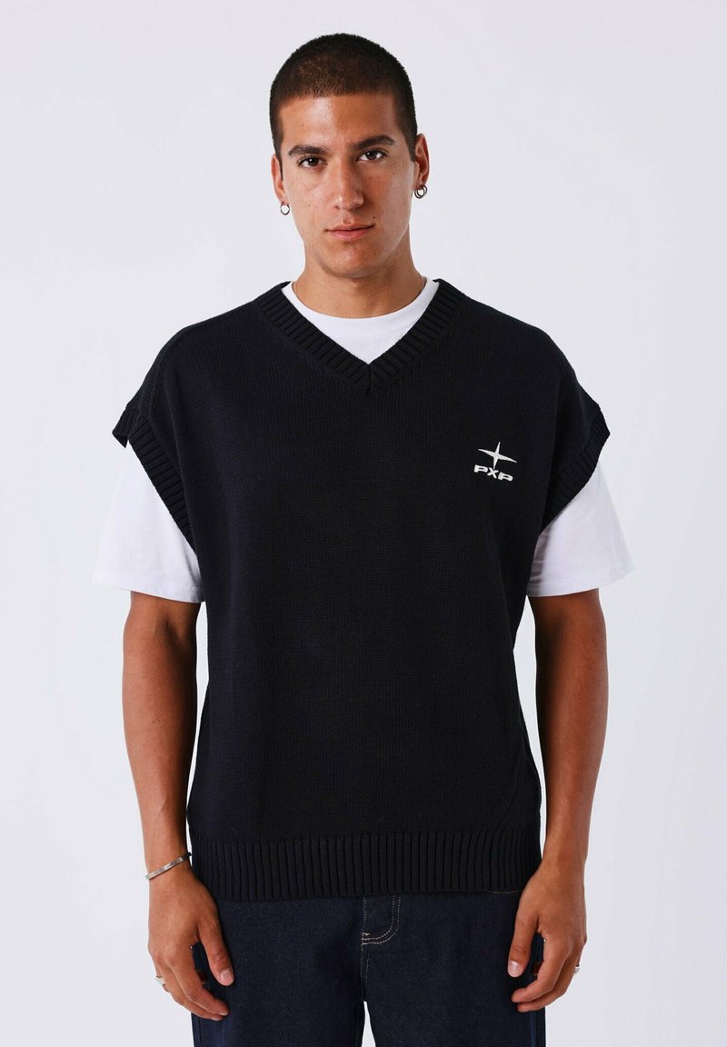 Chaleco de punto negro con escote en V y mangas cortas caídas. Logo blanco en el pecho. Llevado sobre una camiseta blanca.