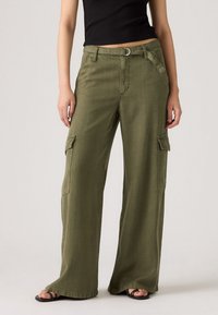 Donna che indossa pantaloni cargo a gamba larga verde oliva con tasche laterali, una cintura di tessuto, un top corto nero e sandali neri con punta aperta.
