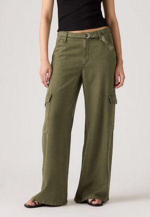 Vrouw gekleed in olijfgroene wijde cargo-broeken met zijzakken, een stoffen riem, een zwarte crop top en zwarte open teeen sandalen.