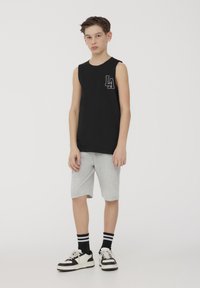 Maglietta nera senza maniche con logo bianco "LA", abbinata a pantaloni corti grigi fino al ginocchio. Il modello indossa scarpe da ginnastica bianche e nere con calze a righe.