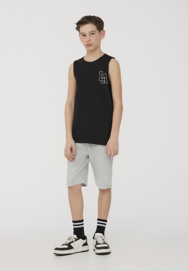 Maglietta nera senza maniche con logo bianco "LA", abbinata a pantaloni corti grigi fino al ginocchio. Il modello indossa scarpe da ginnastica bianche e nere con calze a righe.