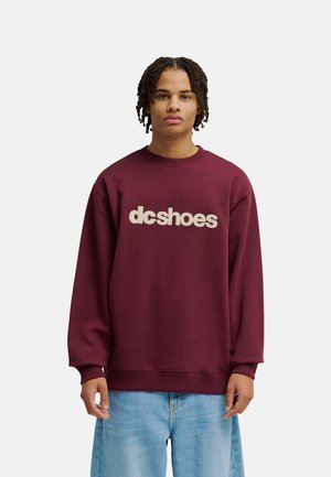 Person steht frontal, trägt ein bordeauxfarbenes dcshoes Sweatshirt und hellblaue Jeans vor einem schlichten, hellen Hintergrund.